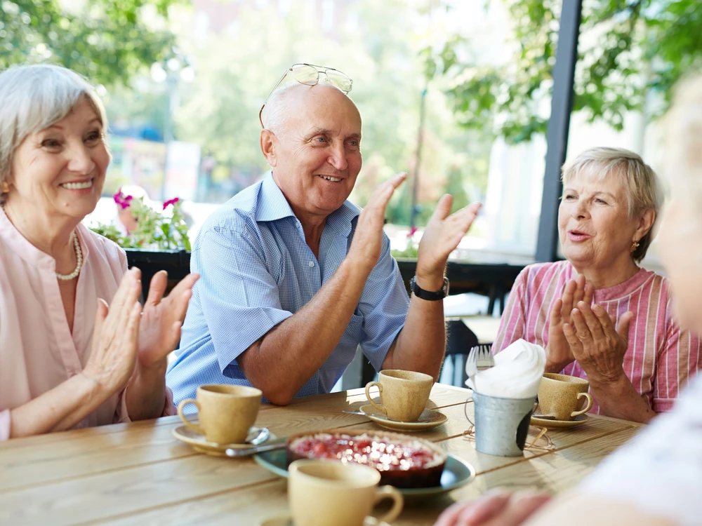 Group of smiling elderly people gathered together in outdoor cafe for tea-drinking and applauding joyfully