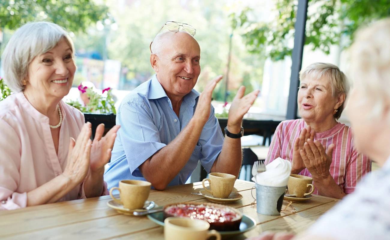 Group of smiling elderly people gathered together in outdoor cafe for tea-drinking and applauding joyfully